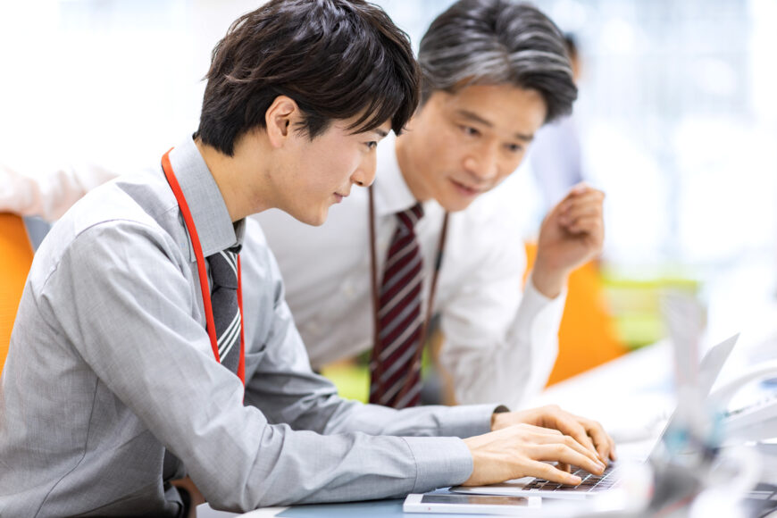 笑顔でPCに向かう若手社員とサポートする上司